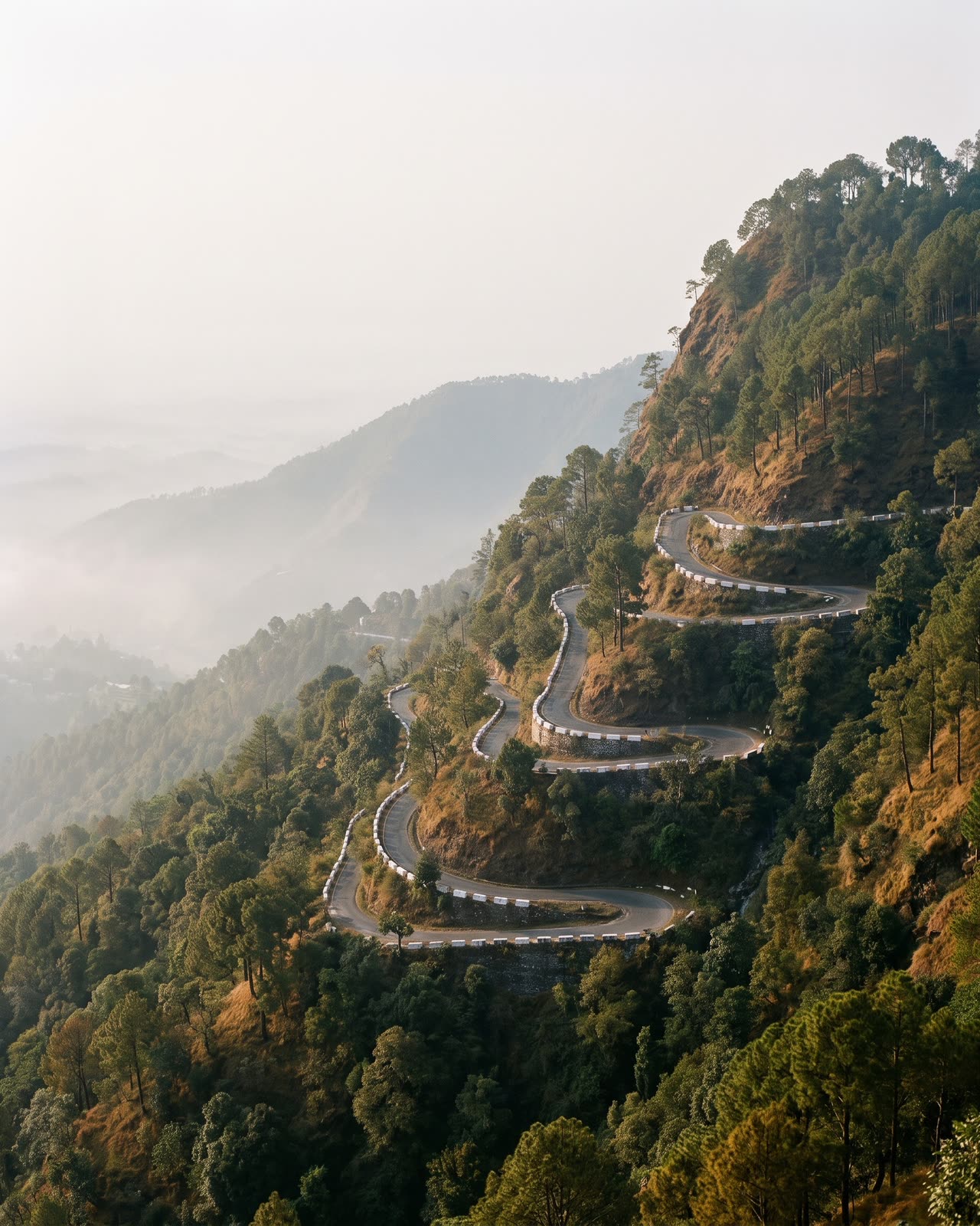 An empty road of tight hairpin bends climbing a forested hillside above a hazy valley.